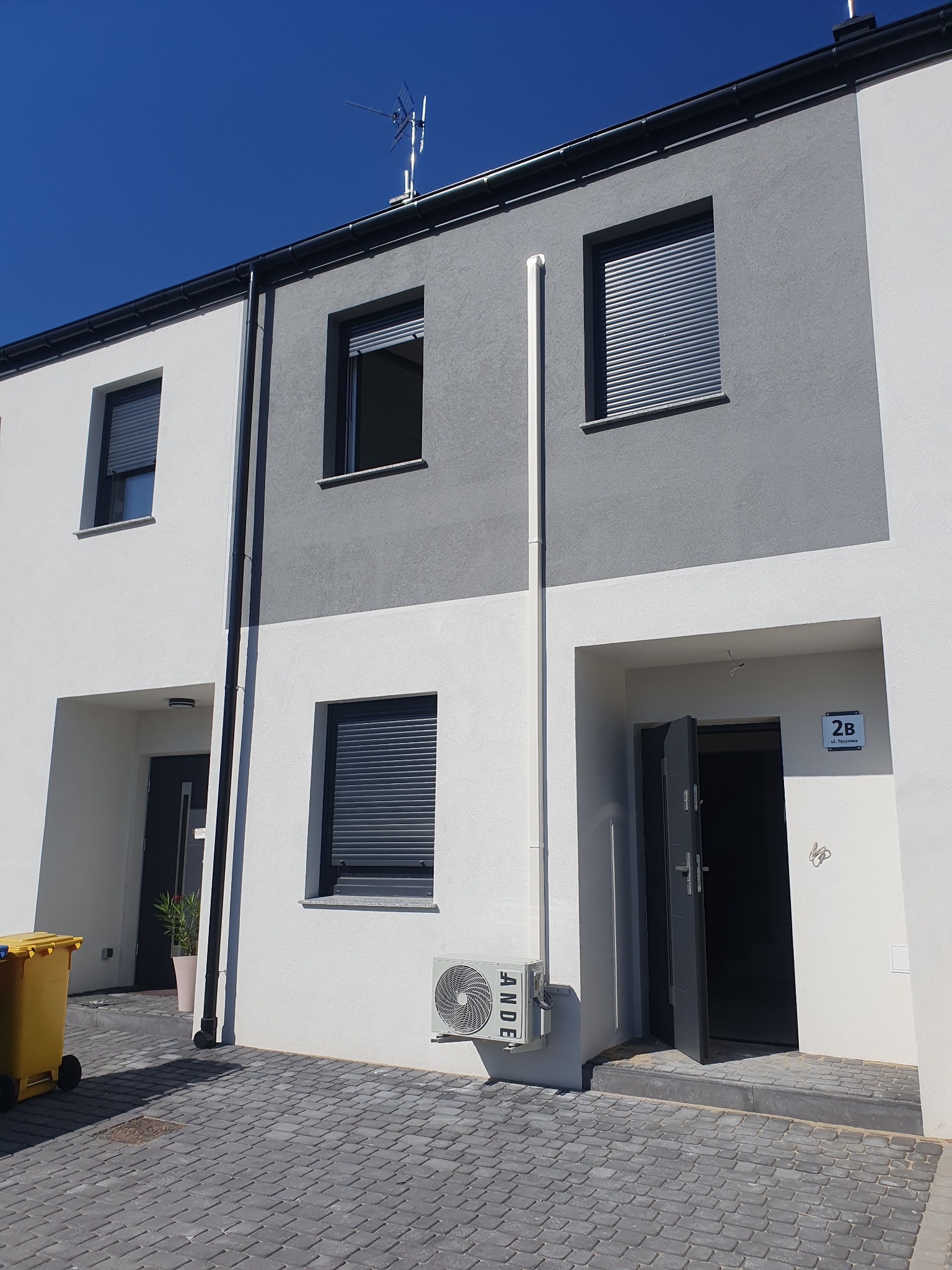 Biało-szary segment bliźniaka z zamontowaną klimatyzacją na ścianie, widoczne rolety zewnętrzne w oknach i żółty kosz na śmieci z boku.