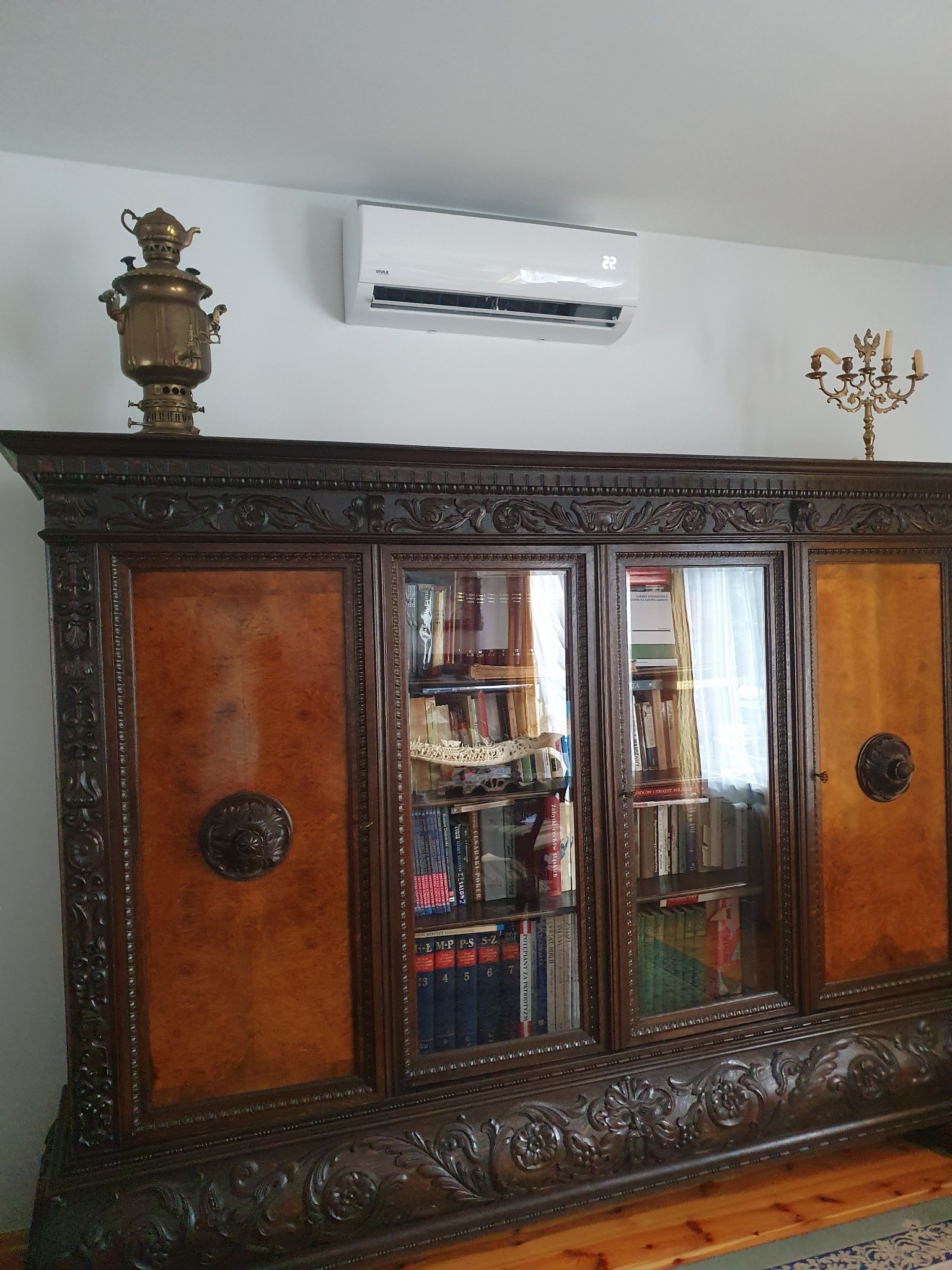 Wnętrze pokoju z antycznym, rzeźbionym kredensem, na którym stoi samowar i świecznik, a nad nim zamontowany klimatyzator ścienny z wyświetlaczem temperatury.