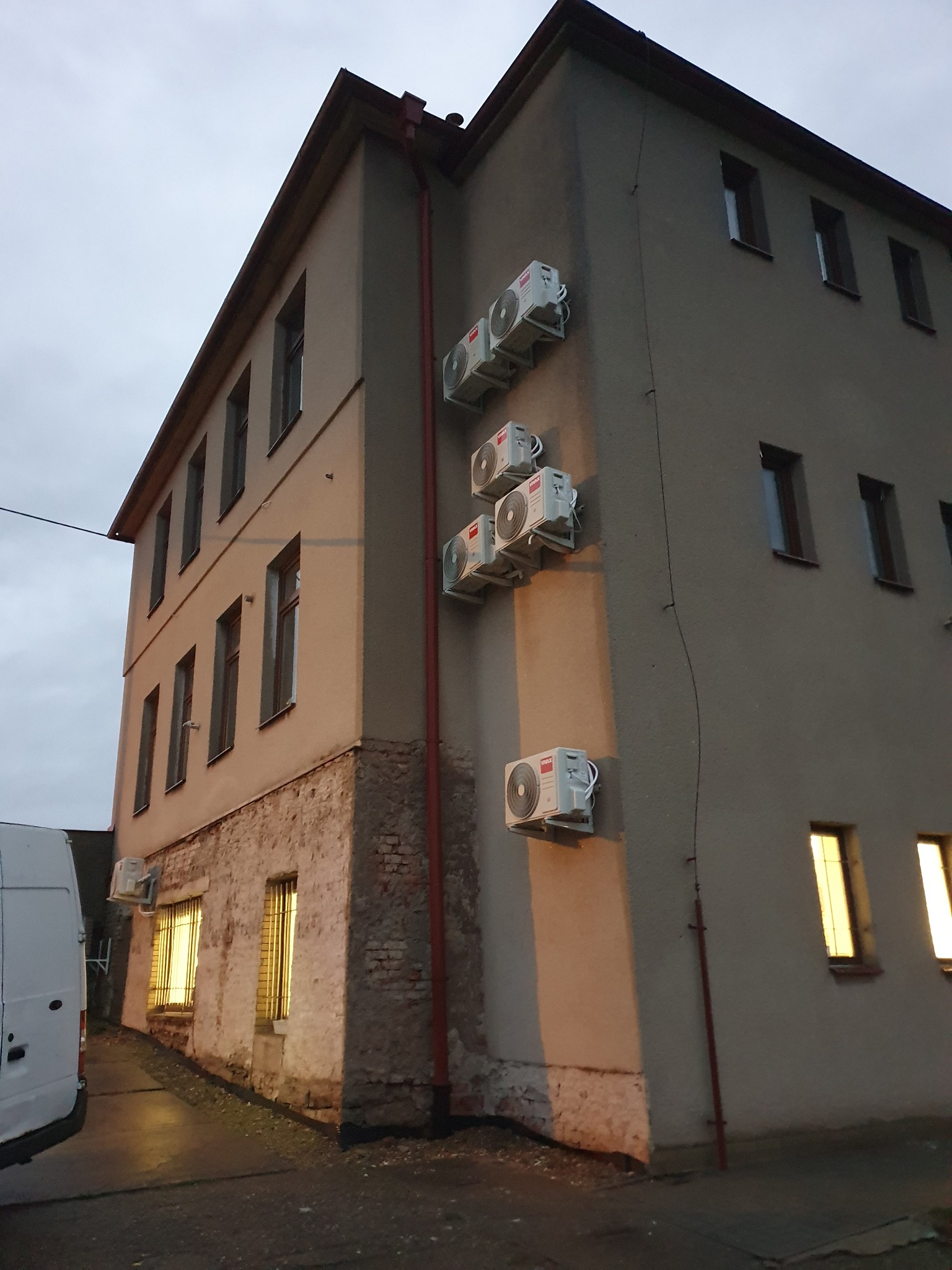 Szereg klimatyzatorów zamontowanych na zewnętrznej ścianie dwupiętrowego budynku biurowego z widocznym fragmentem białego samochodu dostawczego po lewej stronie.