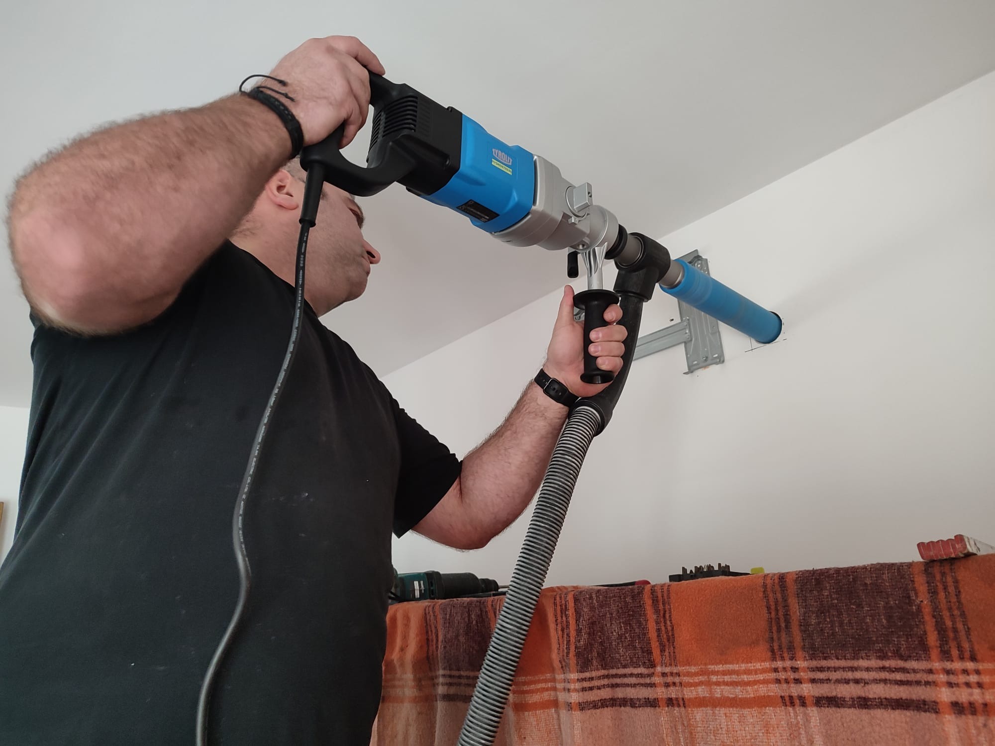Mężczyzna wierci otwór w ścianie pod klimatyzator za pomocą wiertnicy z odsysaniem pyłu, widoczny uchwyt montażowy oraz fragment pomarańczowej tkaniny.