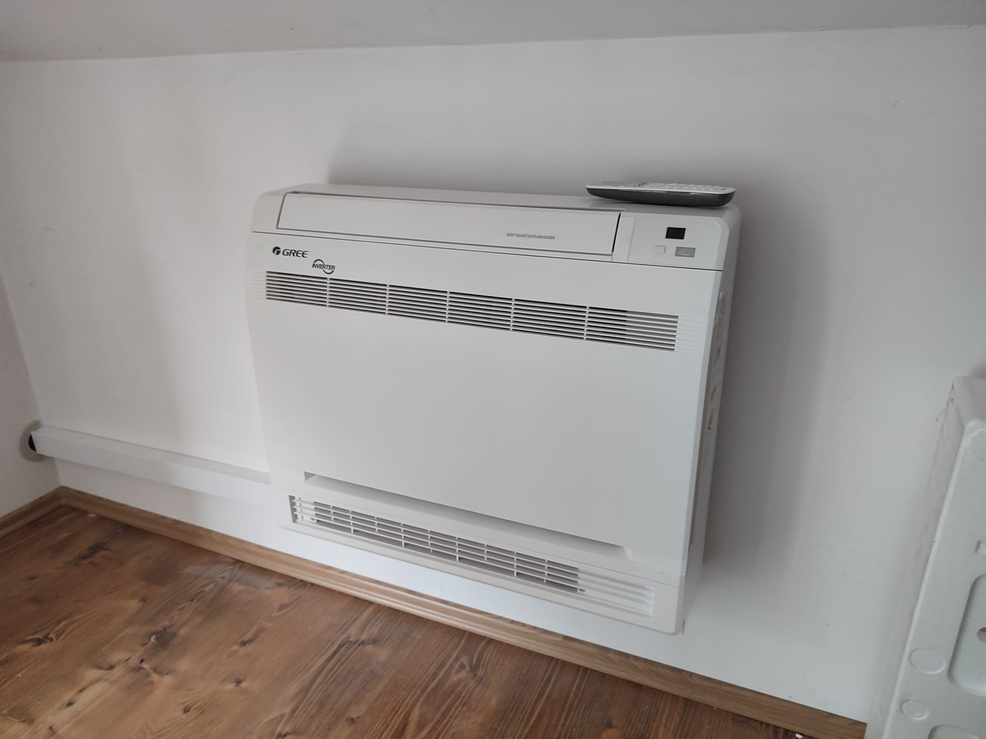 Biały klimatyzator podłogowy marki Gree, zamontowany przy ścianie, z pilotem leżącym na górze, widoczny parkiet na podłodze i listwa przypodłogowa.