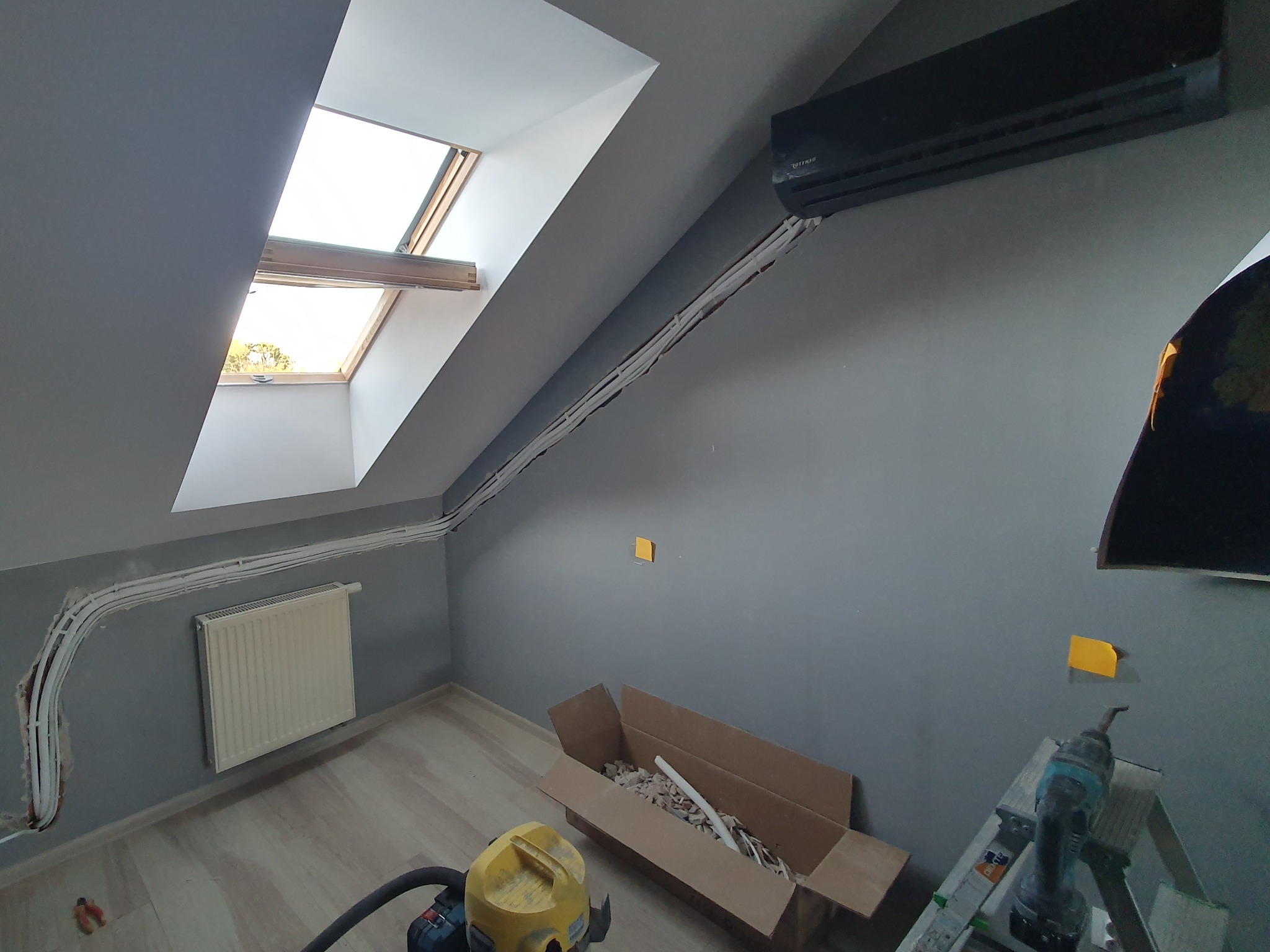 Montaż klimatyzacji w pokoju na poddaszu: widoczny czarny klimatyzator Rotenso na szarej ścianie, białe rurki instalacyjne prowadzone wzdłuż ściany i sufitu, okno dachowe w tle, drabina i narzędzia...