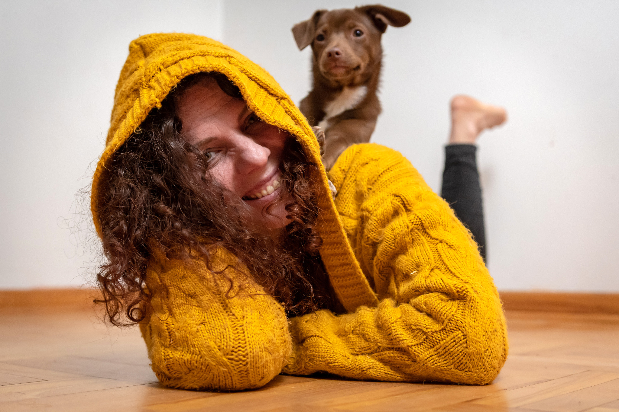 Uśmiechnięta kobieta w musztardowym swetrze z kapturem leży na podłodze, na jej plecach siedzi brązowy szczeniak.