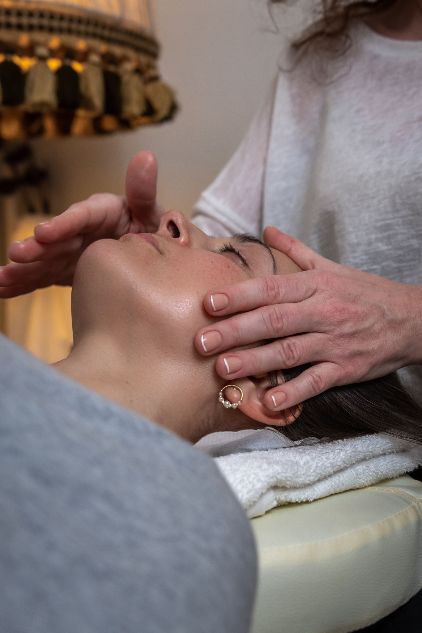 Kobieta podczas masażu twarzy, dłonie masażysty delikatnie dotykają jej czoła i policzków, widoczne złote kolczyki z perełkami.