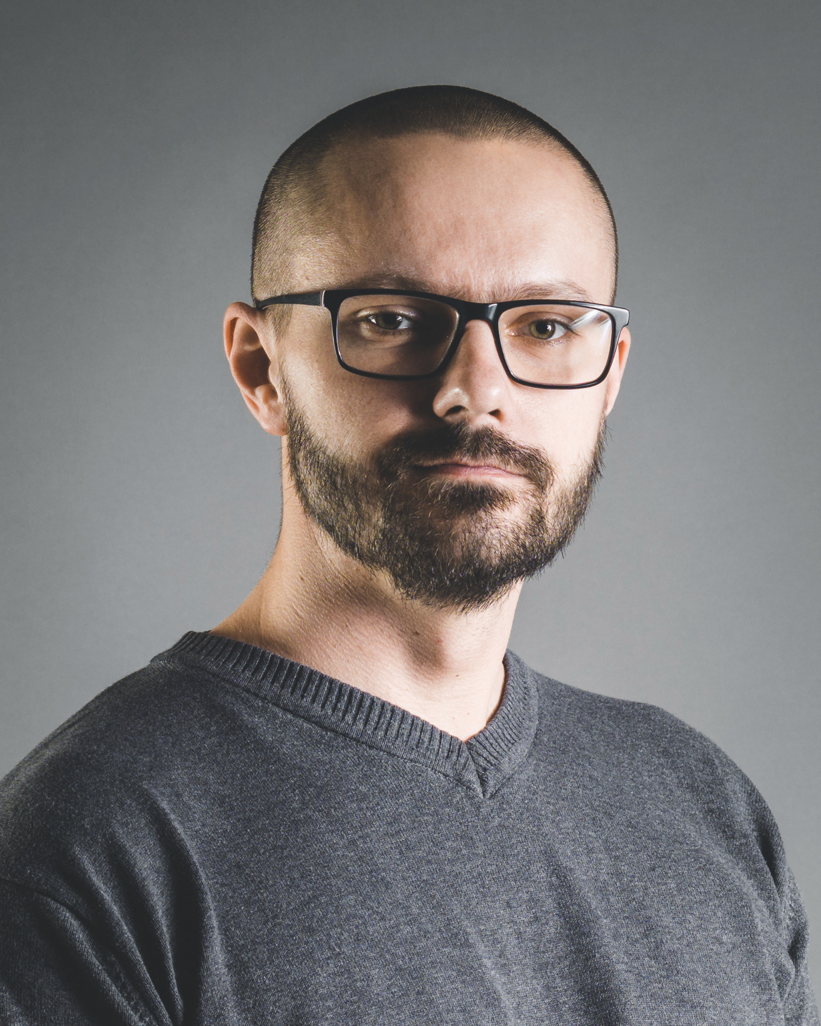 Portret mężczyzny z brodą i okularami na szarym tle. Ujęcie od przodu, skupione na twarzy. Neutralny wyraz twarzy, krótko ostrzyżone włosy.