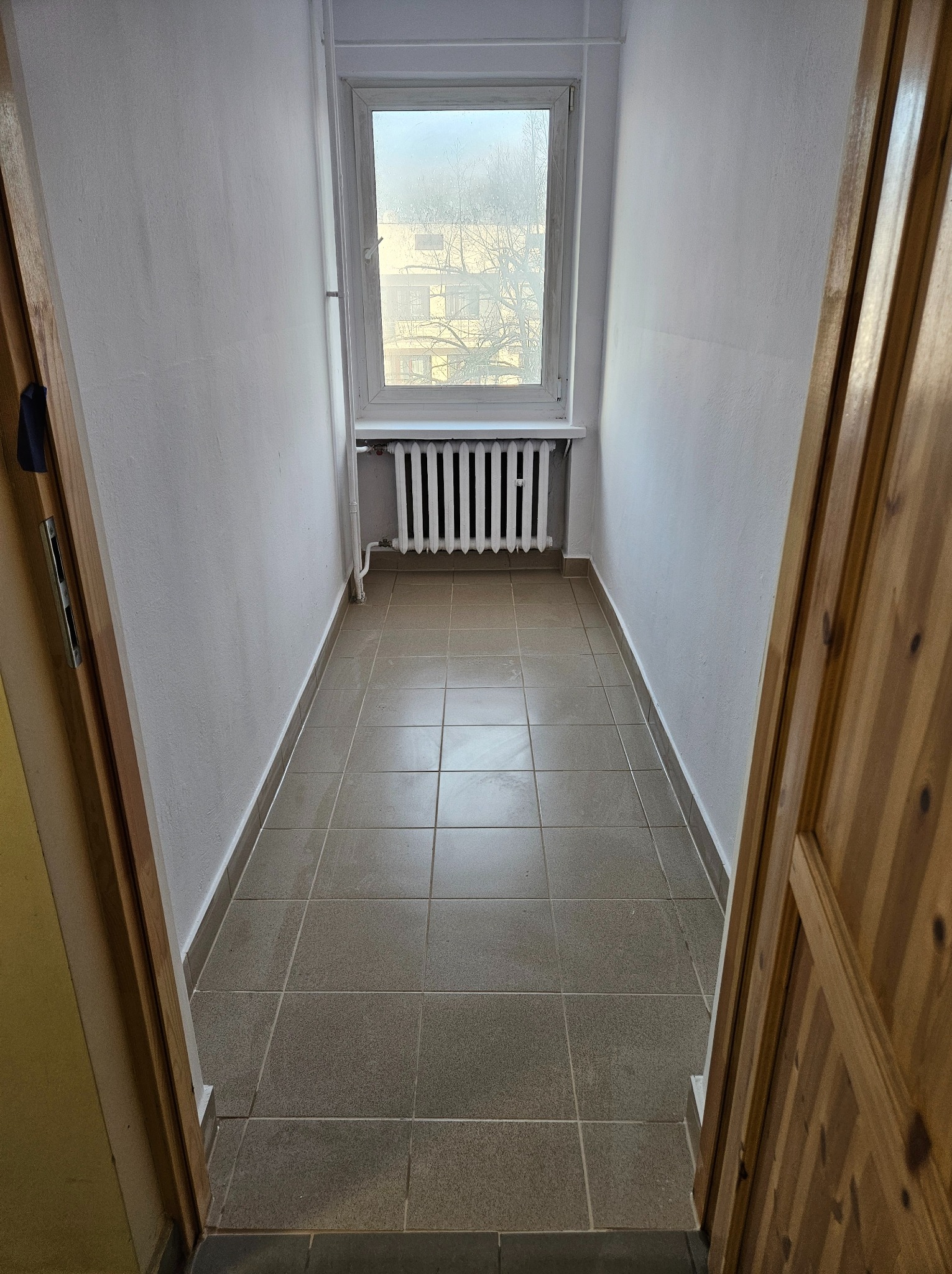 Wąskie pomieszczenie suszarni po odświeżeniu, z białymi ścianami, grzejnikiem pod oknem i szarą terakotą na podłodze, widok zza drewnianych drzwi.