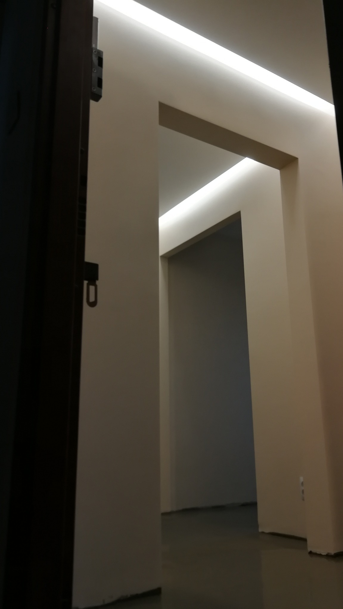 Nowoczesne oświetlenie LED w przedpokoju z podwieszanym sufitem i minimalistycznym wykończeniem ścian w odcieniach beżu, widoczne zza otwartych drzwi wejściowych.