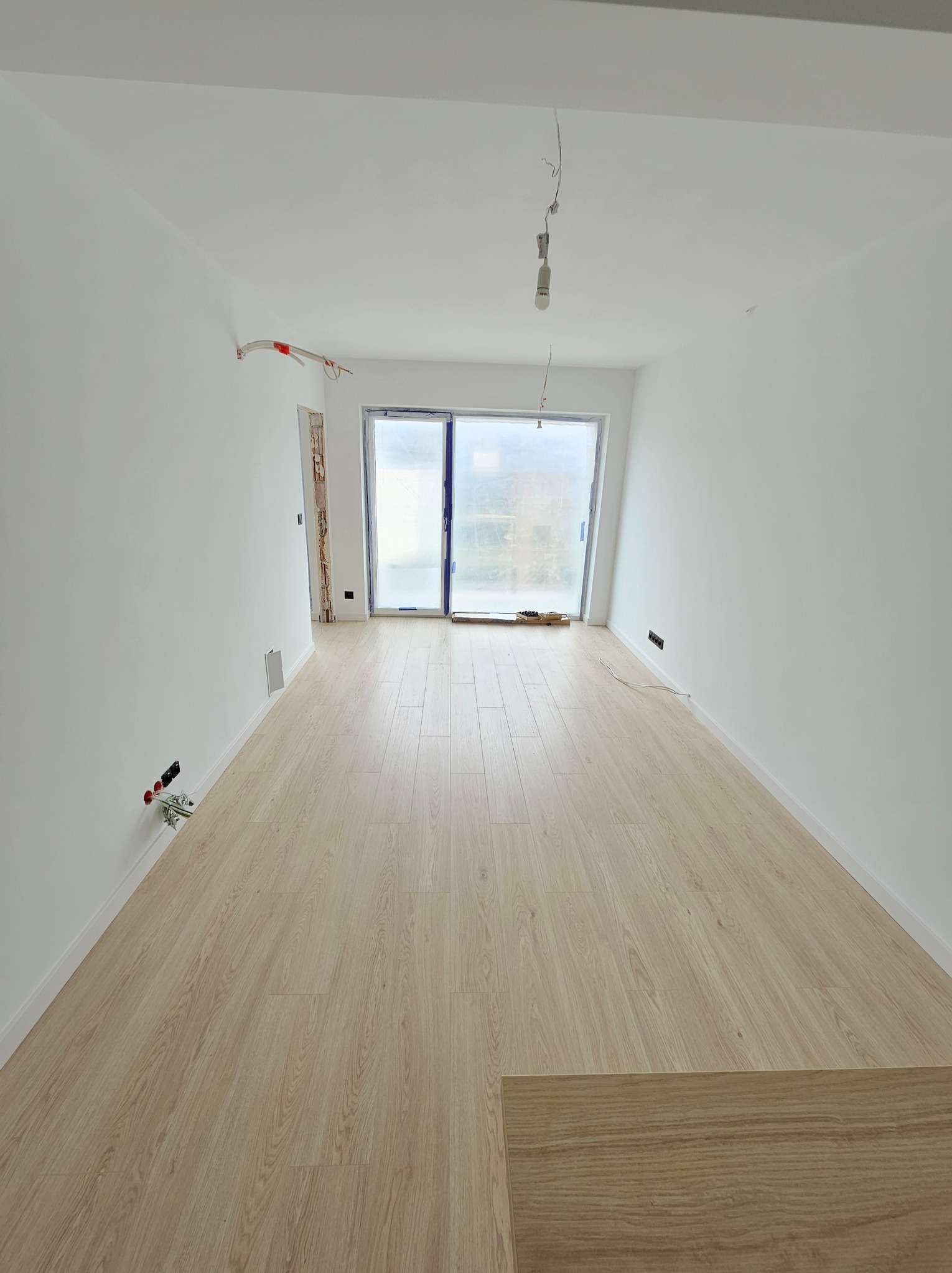 Nowe mieszkanie po remoncie: jasne panele podłogowe, białe ściany z gładzią, widoczne gniazdka elektryczne bez osprzętu i wystające kable sufitowe.