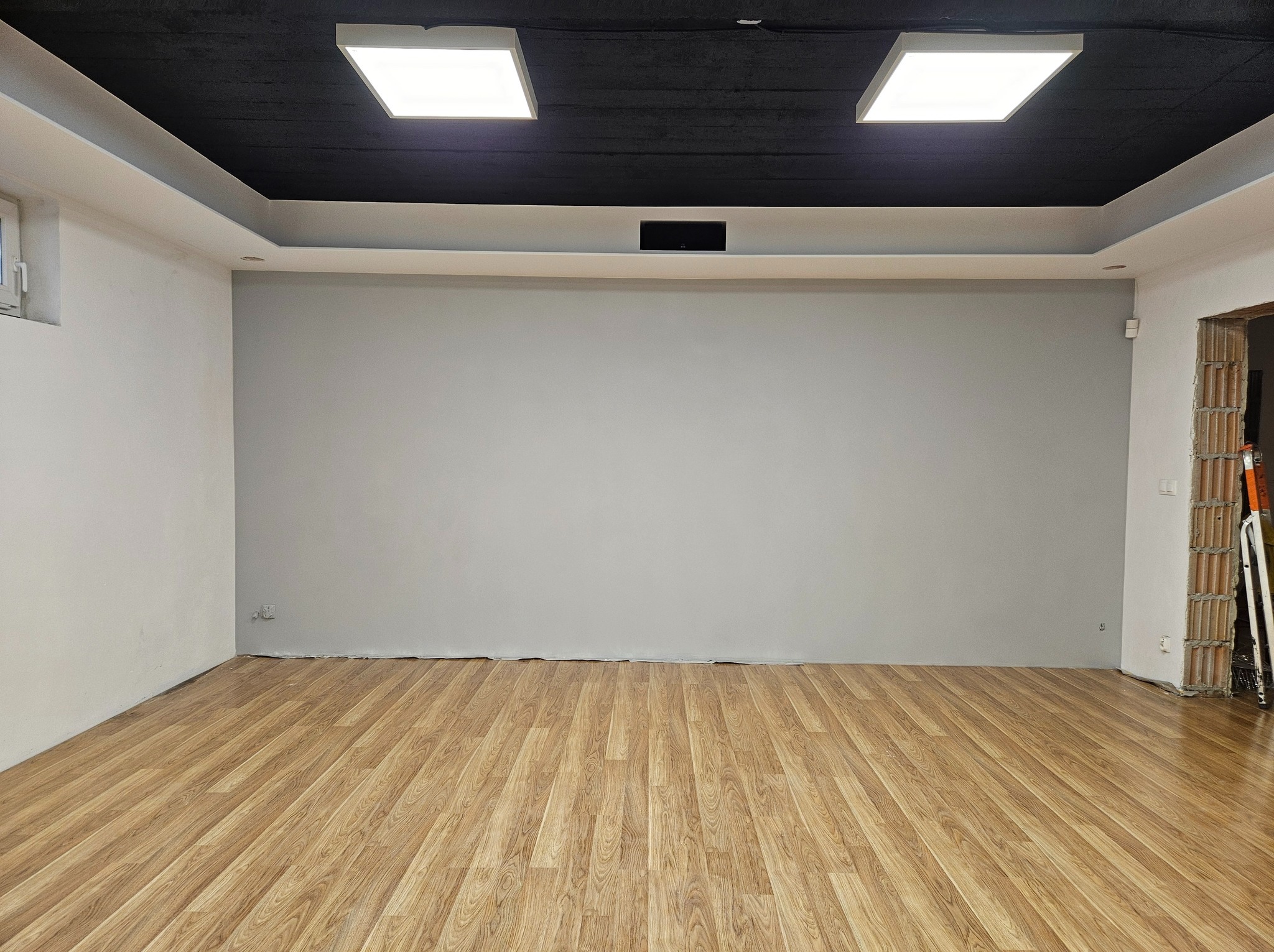 Wykończone wnętrze z panelami podłogowymi, szarą ścianą przygotowaną pod malowanie ekranowe i czarnym sufitem podwieszanym z dwoma kwadratowymi lampami LED.