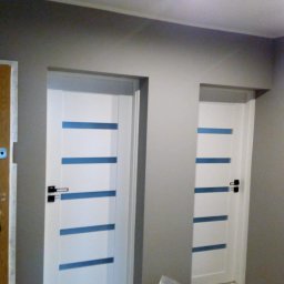 Robert Rutz - Dwa białe, nowoczesne drzwi wewnętrzne z poziomymi, niebieskimi paskami zamontowane w szarym korytarzu, obok brązowe drzwi wejściowe z zamkiem.