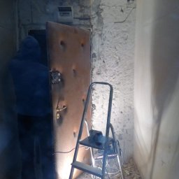 Robert Rutz - Montera drzwi w trakcie pracy: widoczna metalowa ościeżnica, częściowo otynkowana ściana, drabina i gruz na podłodze, oświetlenie robocze.