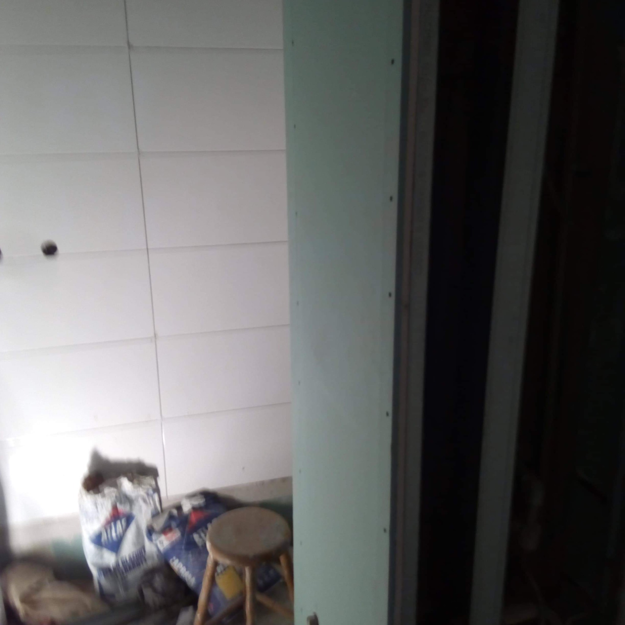 Ściana z białych płytek w łazience w trakcie remontu, widoczny worek zaprawy i taboret oraz fragment ściany z płyty kartonowo-gipsowej.