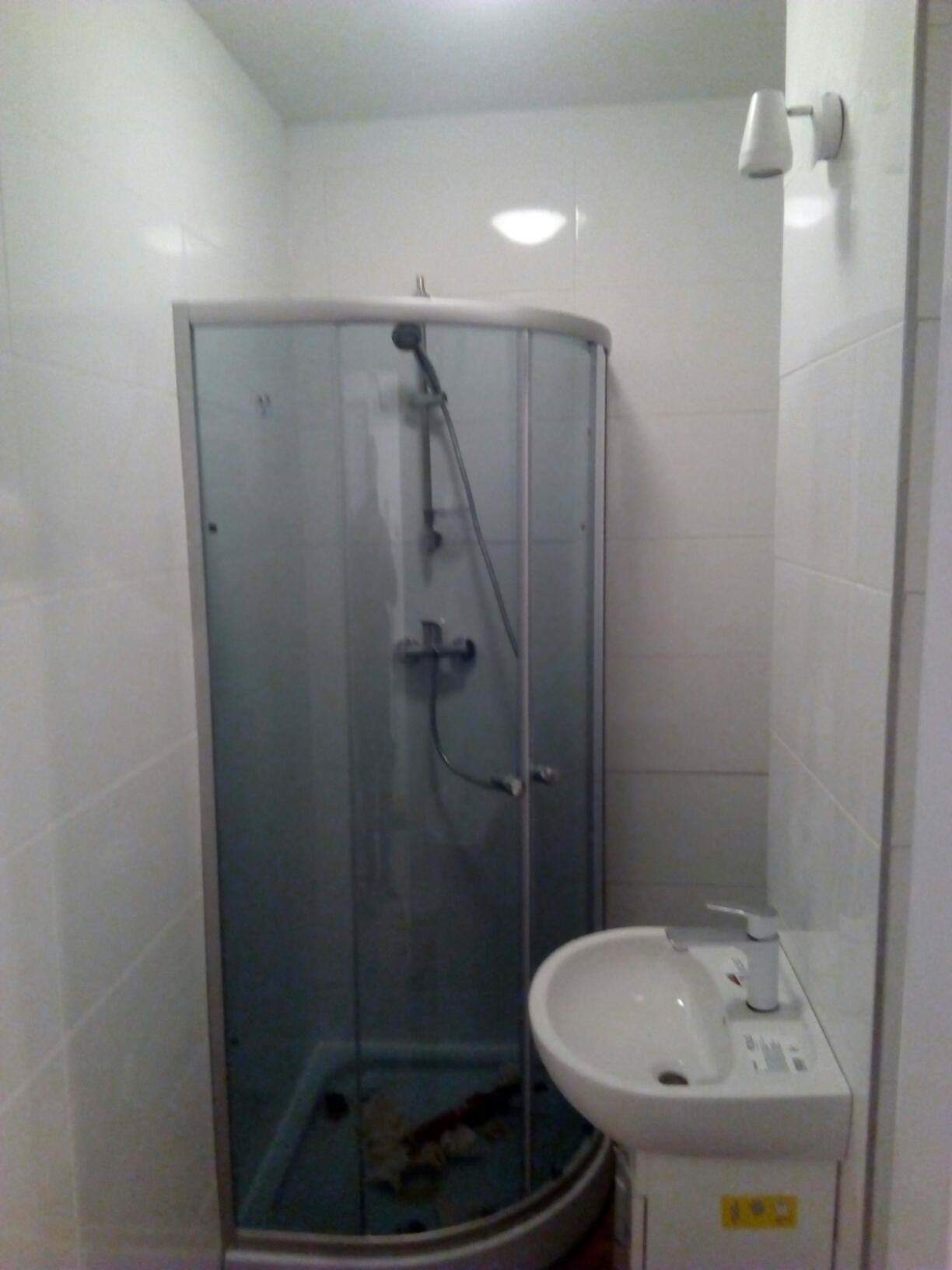 Narożna kabina prysznicowa z matowego szkła i biała umywalka w małej łazience wyłożonej białymi płytkami, widoczna lampa ścienna.