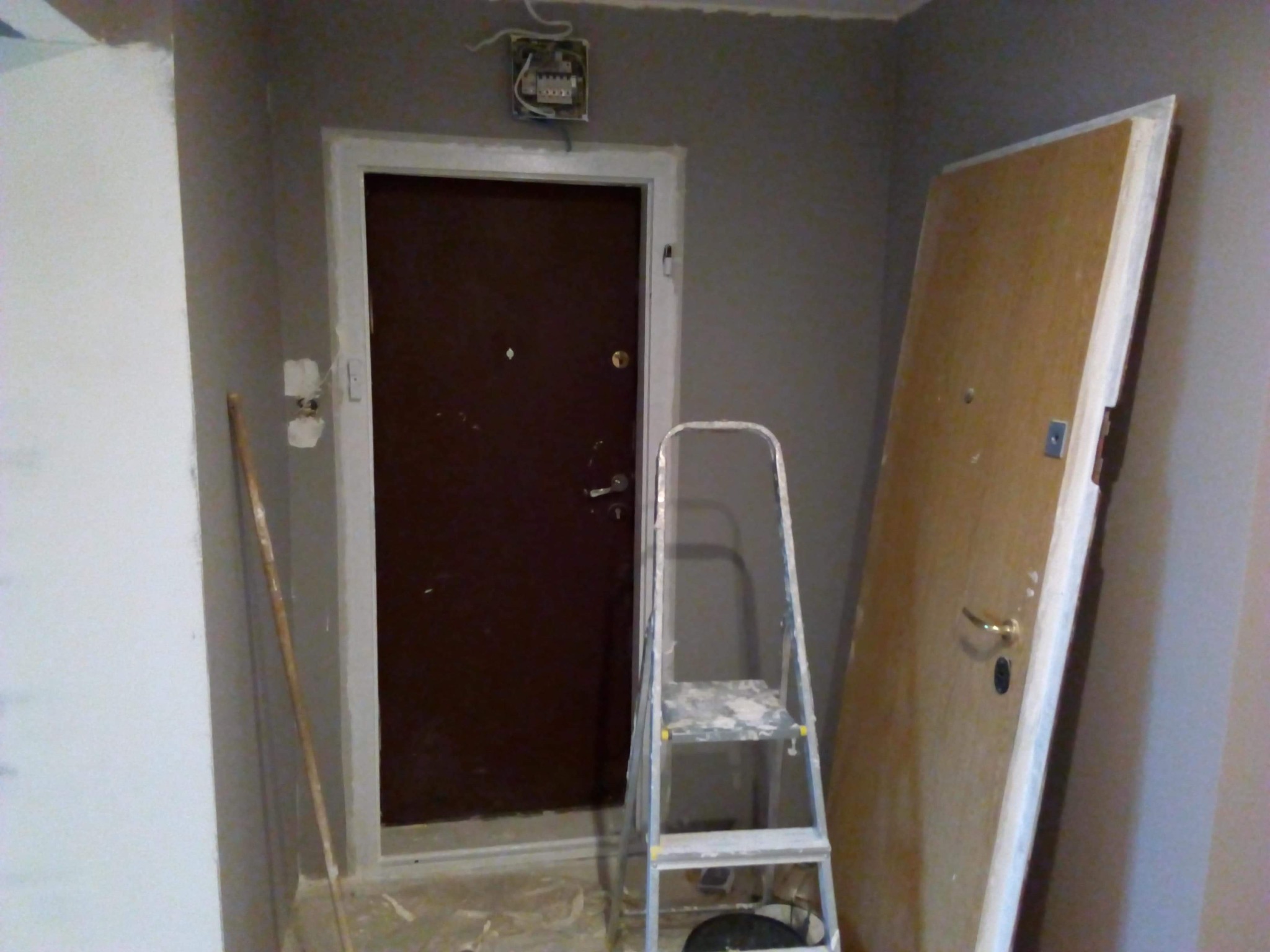 Wnętrze w trakcie prac wykończeniowych: pomalowane ściany szarą farbą, widoczna skrzynka elektryczna, brązowe drzwi wejściowe z ościeżnicą przygotowaną do malowania, drabina malarska i drzwi...