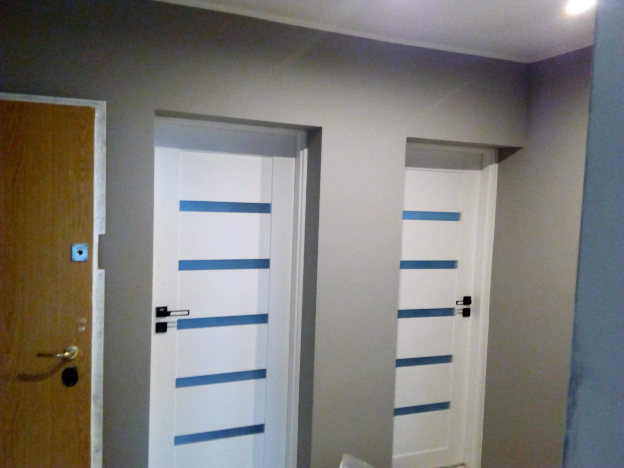 Dwa białe, nowoczesne drzwi wewnętrzne z poziomymi, niebieskimi paskami zamontowane w szarym korytarzu, obok brązowe drzwi wejściowe z zamkiem.