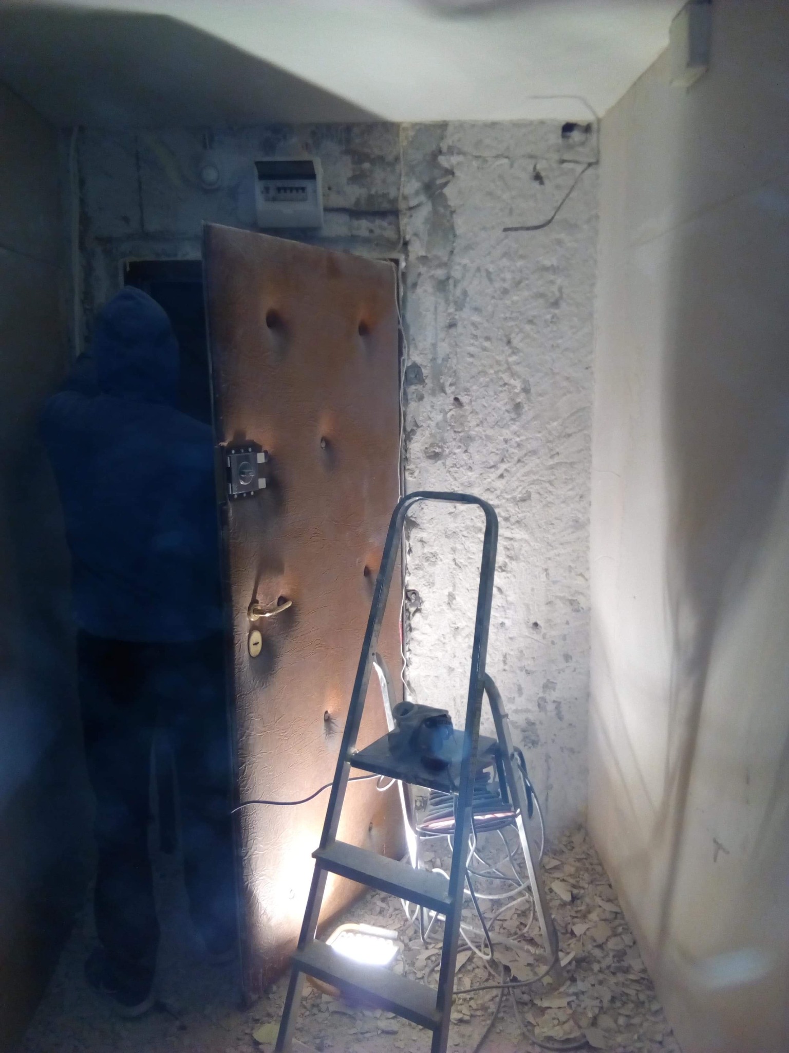 Montera drzwi w trakcie pracy: widoczna metalowa ościeżnica, częściowo otynkowana ściana, drabina i gruz na podłodze, oświetlenie robocze.