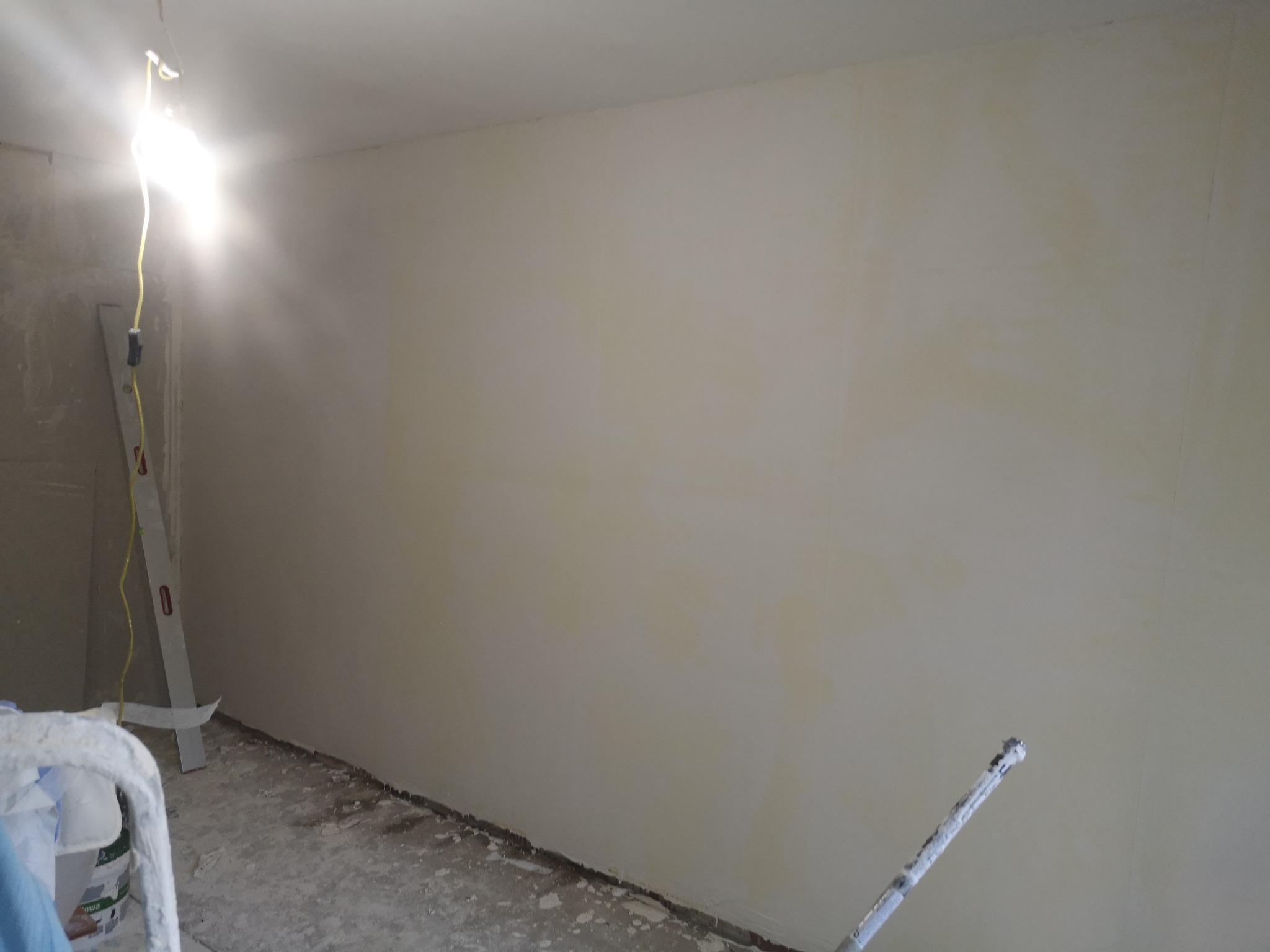 Ściana w trakcie malowania na jasny odcień, widoczna drabina i wiadro z farbą, surowa podłoga, oświetlenie halogenowe.