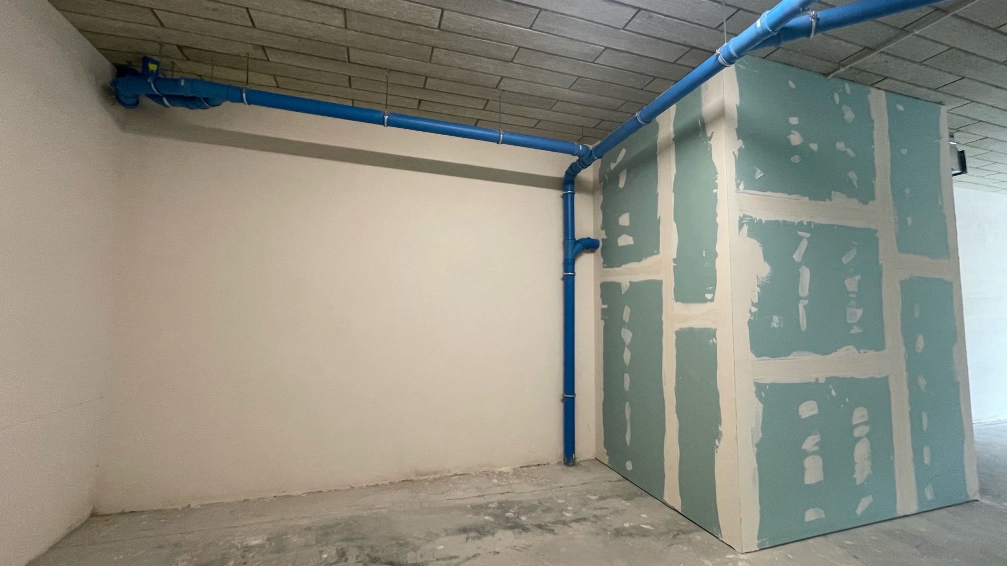 Wnętrze w trakcie prac wykończeniowych: ściana z płyt kartonowo-gipsowych, niebieskie rury instalacyjne pod stropem, surowa podłoga.