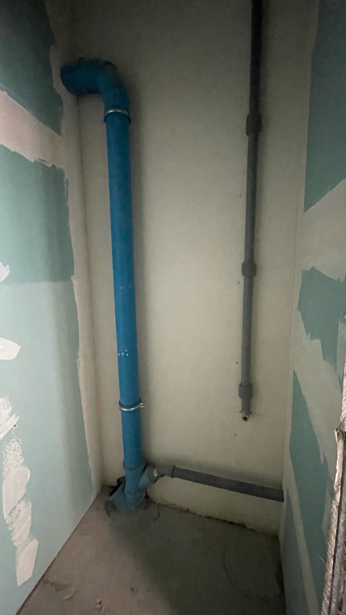 Niebieska rura kanalizacyjna z obejmami mocowana do ściany w rogu pomieszczenia, widoczny fragment szarej rury poziomej i pionowej, ściany w trakcie szpachlowania płyt kartonowo-gipsowych.