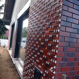 OLEH SHPAKOVSKYI - Kątowe ujęcie fragmentu elewacji budynku w trakcie układania klinkierowej okładziny z użyciem krzyżyków dystansowych, widoczny fragment okna i otoczenie budynku.