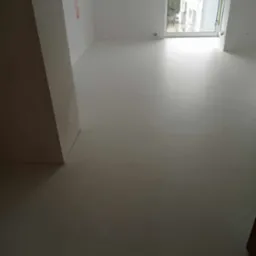 Jasne, puste pomieszczenie z białymi ścianami i podłogą w odcieniach szarości, duże okno z widokiem na budynki na zewnątrz, widoczny fragment futryny drzwi po prawej stronie kadru.