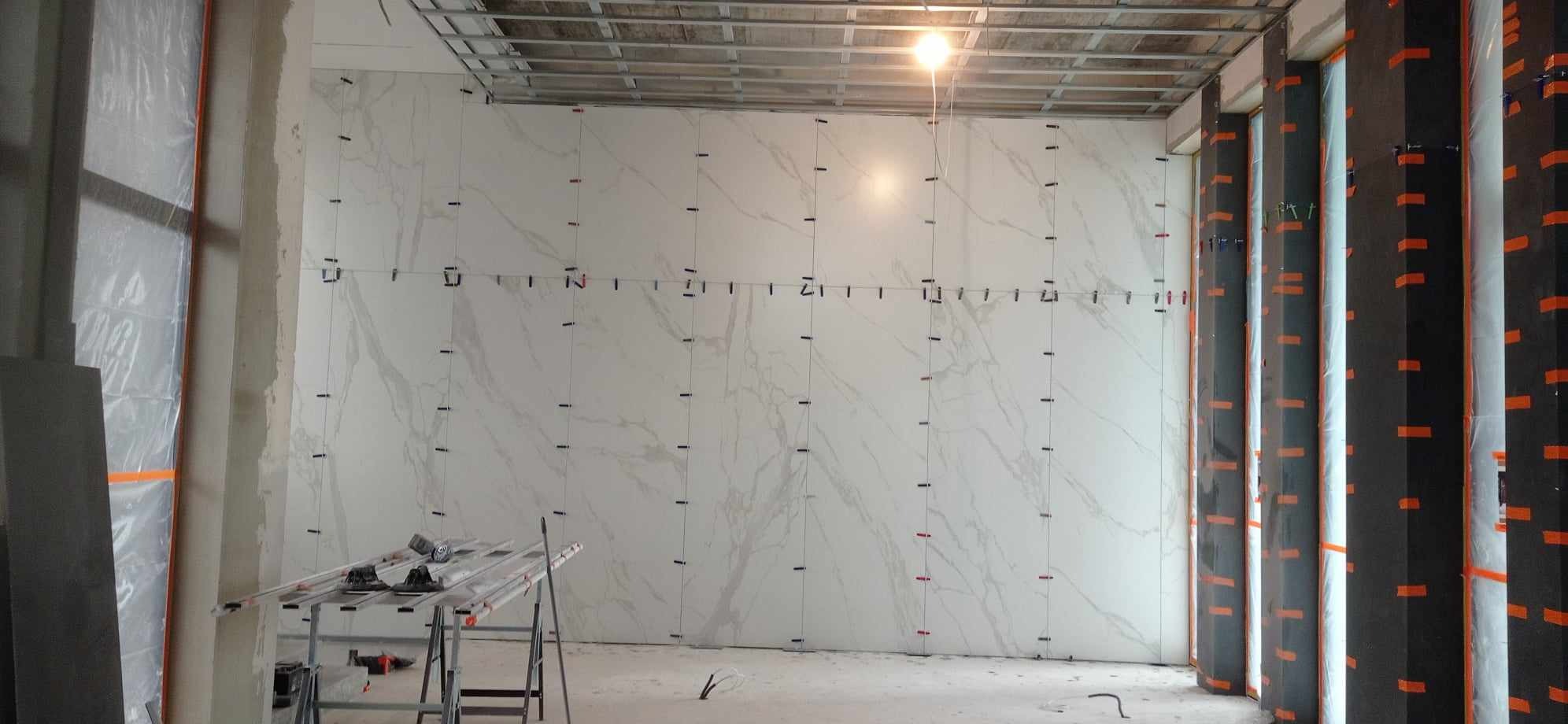 Ściana w trakcie montażu wielkoformatowych płyt imitujących marmur, widoczne systemy poziomowania płytek, metalowa konstrukcja sufitu podwieszanego oraz zabezpieczone folią i taśmą narożniki ścian.