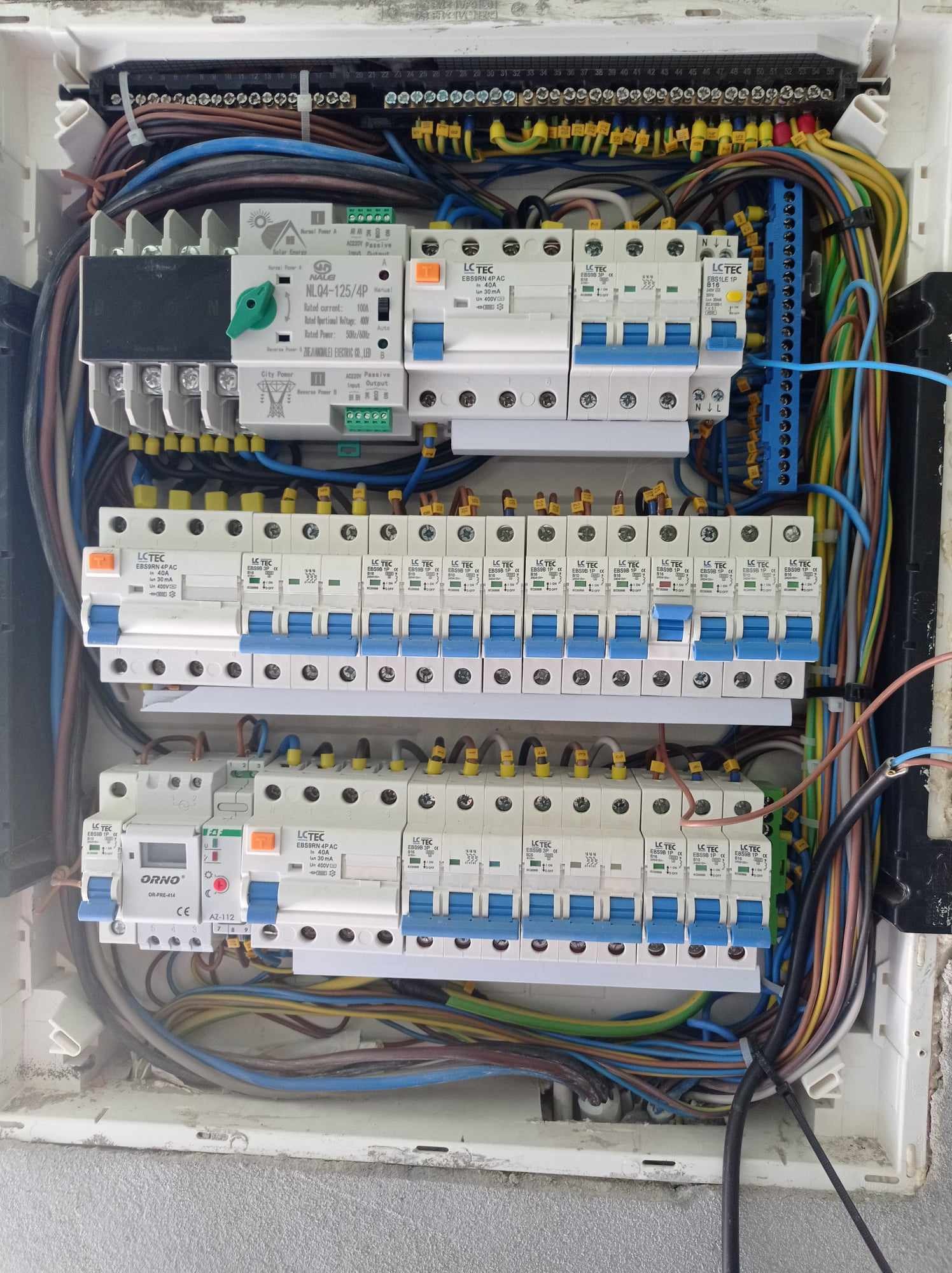 Rozdzielnica elektryczna z licznymi zabezpieczeniami, przewodami i listwami zaciskowymi, widoczne elementy sterujące i pomiarowe, zainstalowana w białej obudowie.