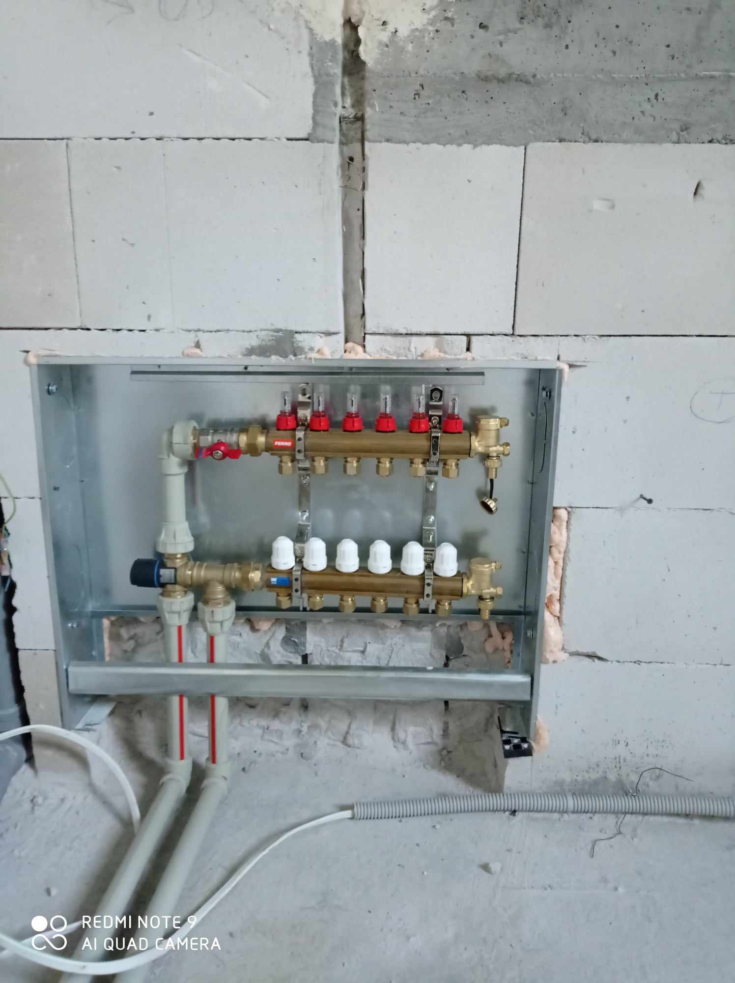 Rozdzielacz ogrzewania podłogowego zamontowany w metalowej skrzynce, widoczne rury doprowadzające i odprowadzające wodę, ściana z białych bloczków betonowych w stanie surowym.