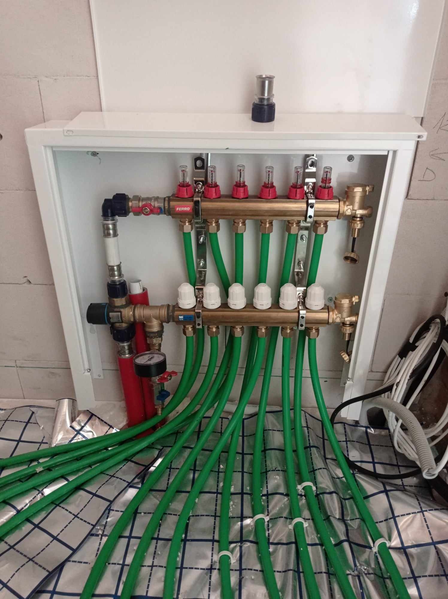 Rozdzielacz ogrzewania podłogowego FERRO w białej szafce, z zielonymi rurami na folii izolacyjnej z nadrukiem kratki.
