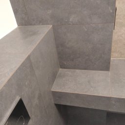 Serhii Liashko SL-BUD - Szare, matowe płytki gresowe w łazience, ułożone w narożniku z półką, widoczny fragment zabudowy stelaża WC.