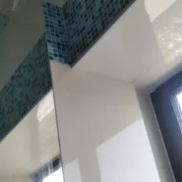 Fragment wykończonej łazienki z mozaiką w odcieniach niebieskiego i turkusu, ułożoną wzdłuż górnej krawędzi ściany i nad oknem, z białymi, błyszczącymi płytami poniżej.