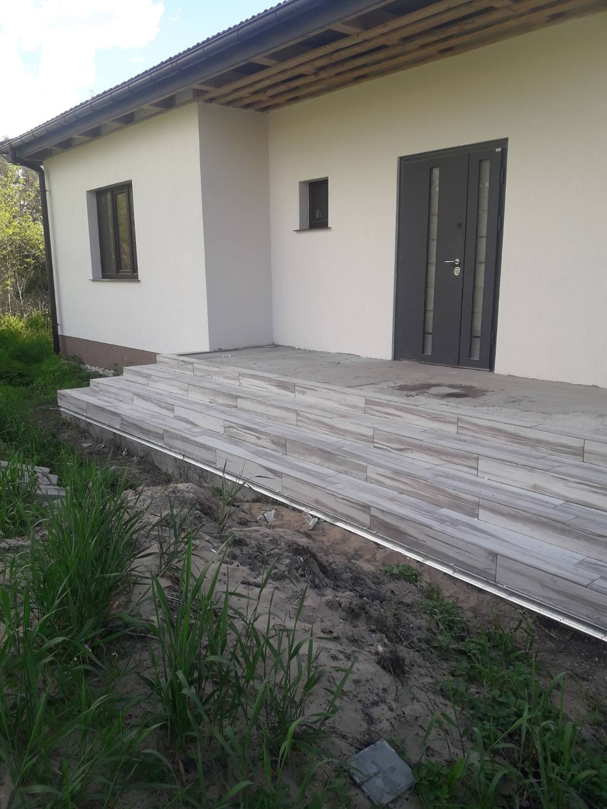 Wyłożone płytkami schody wejściowe do domu z szarą stolarką okienną i drzwiową, widoczne trawy i ziemia w otoczeniu.