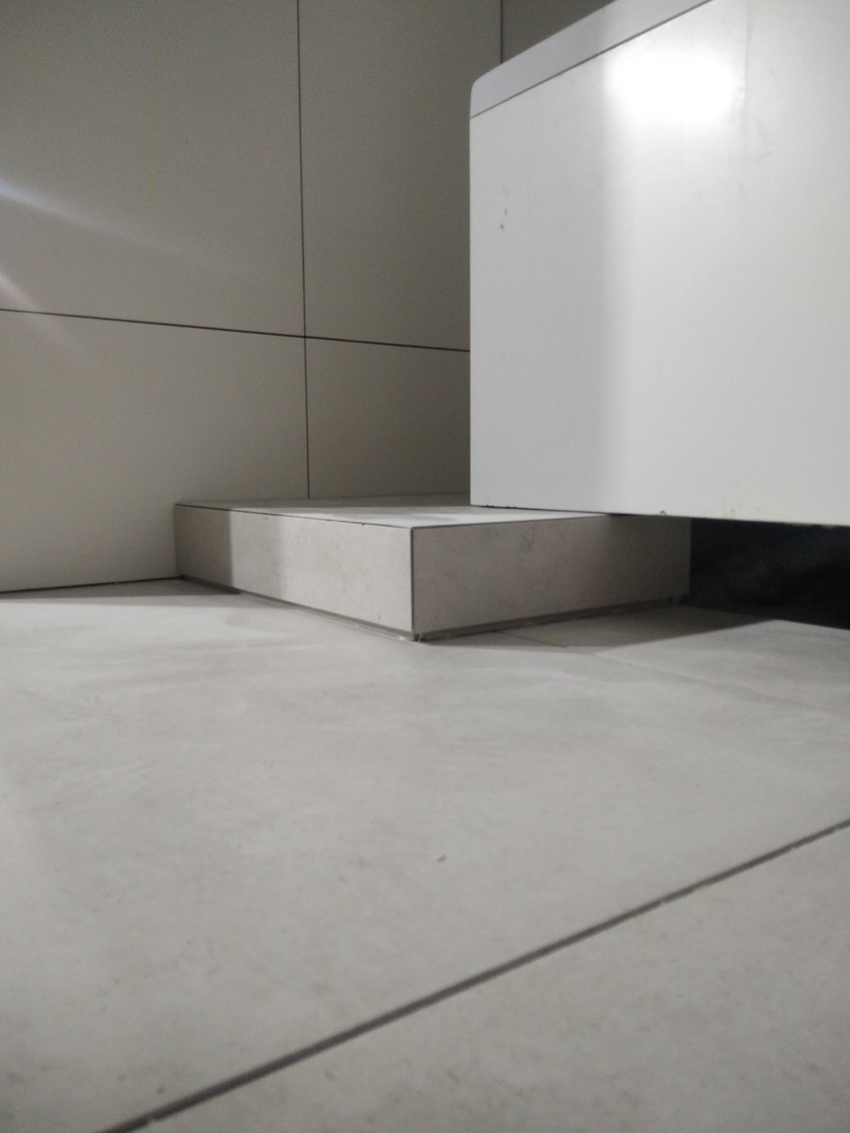 Szara, minimalistyczna łazienka z płytkami podłogowymi i zabudową pod umywalką, widoczne fugi i narożniki.