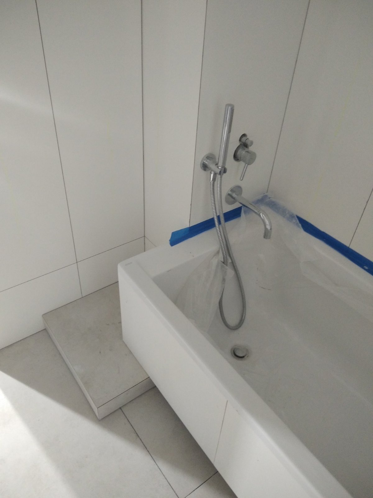 Biała, prostokątna wanna zabezpieczona folią i niebieską taśmą malarską podczas prac wykończeniowych w łazience w Toruniu, widoczne białe płytki na ścianach i chromowana bateria.