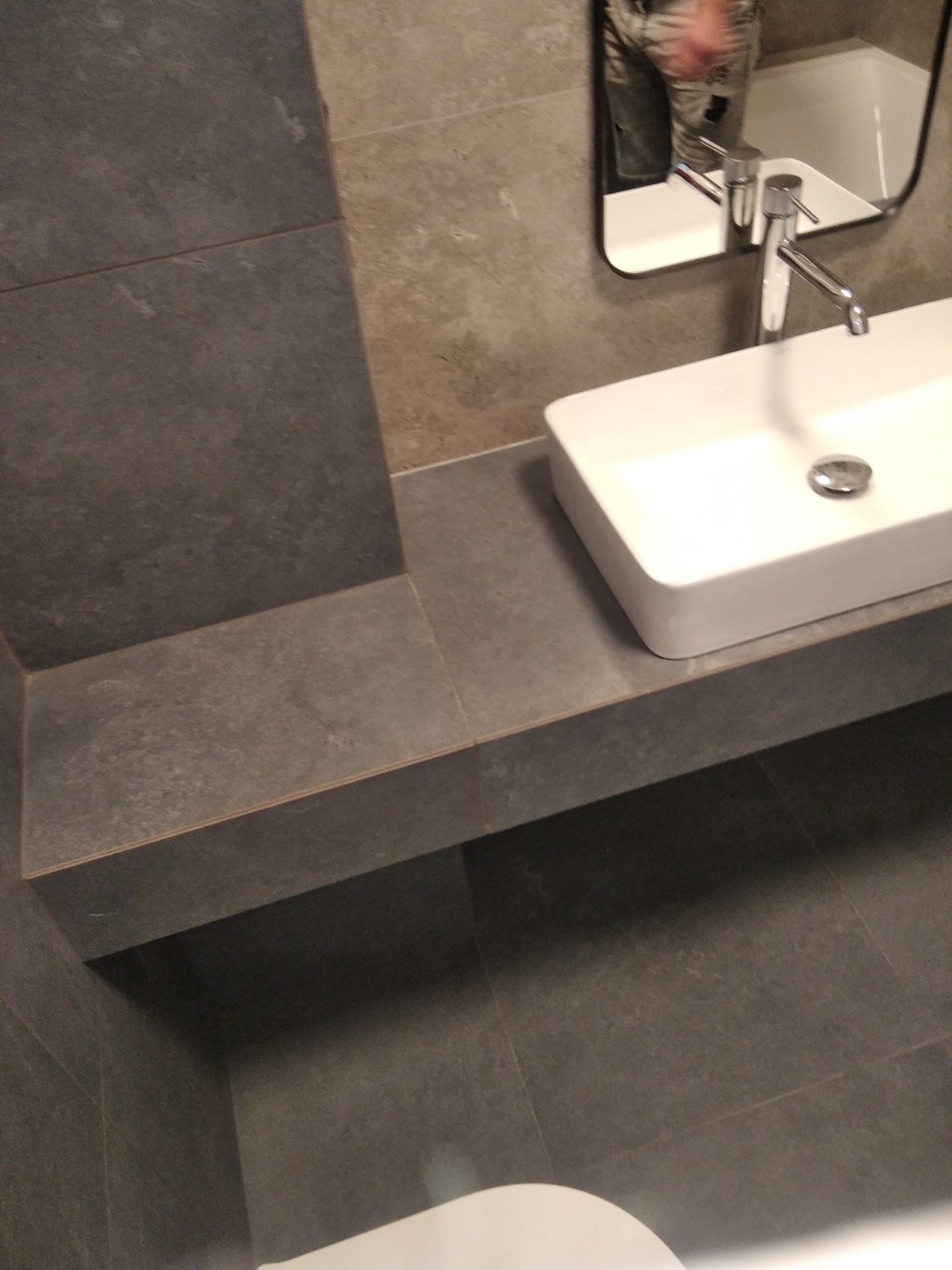 Nowoczesna łazienka z prostokątną umywalką nablatową, chromowaną baterią i szarą płytką ceramiczną na ścianach i podłodze, odbicie osoby w lustrze.