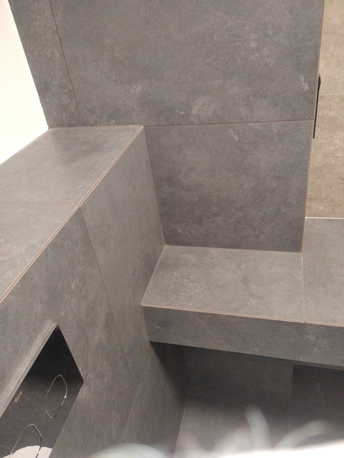 Szare, matowe płytki gresowe w łazience, ułożone w narożniku z półką, widoczny fragment zabudowy stelaża WC.