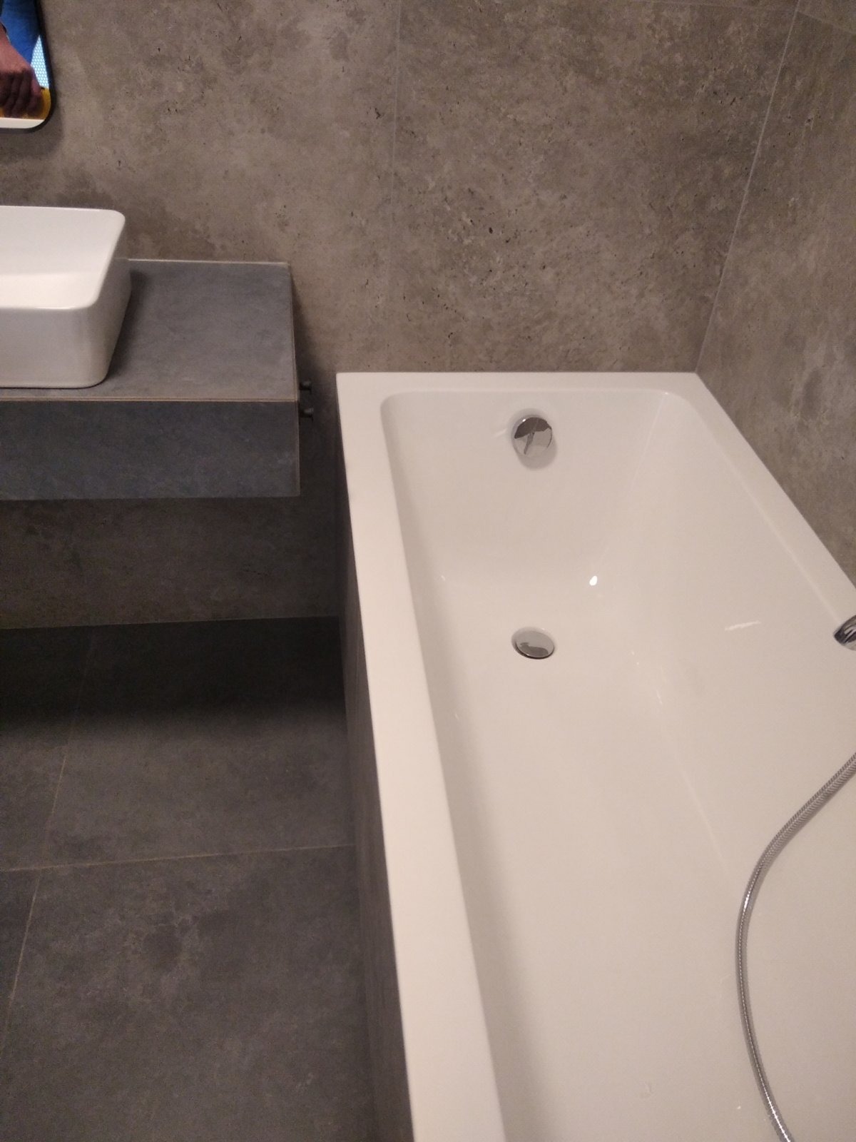 Nowoczesna łazienka z białą wanną, szarą umywalką i płytkami imitującymi beton na ścianach i podłodze.