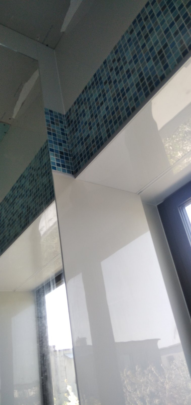 Fragment wykończonej łazienki z mozaiką w odcieniach niebieskiego i turkusu, ułożoną wzdłuż górnej krawędzi ściany i nad oknem, z białymi, błyszczącymi płytami poniżej.