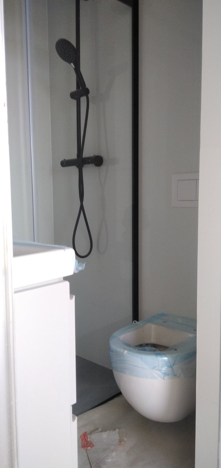 Nowa łazienka w trakcie remontu: czarna bateria prysznicowa z deszczownicą, biała podwieszana toaleta zabezpieczona folią, szare płytki podłogowe, widok zza framugi drzwi.