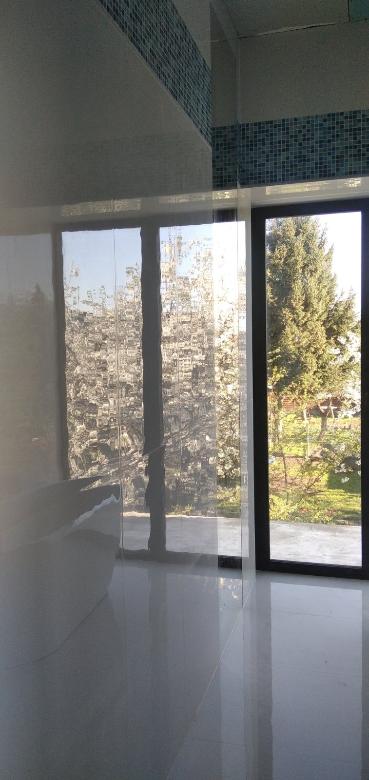 Nowoczesna łazienka z dużym oknem z widokiem na ogród, wykończona błyszczącymi, białymi płytkami podłogowymi i mozaiką w odcieniach niebieskiego pod sufitem.