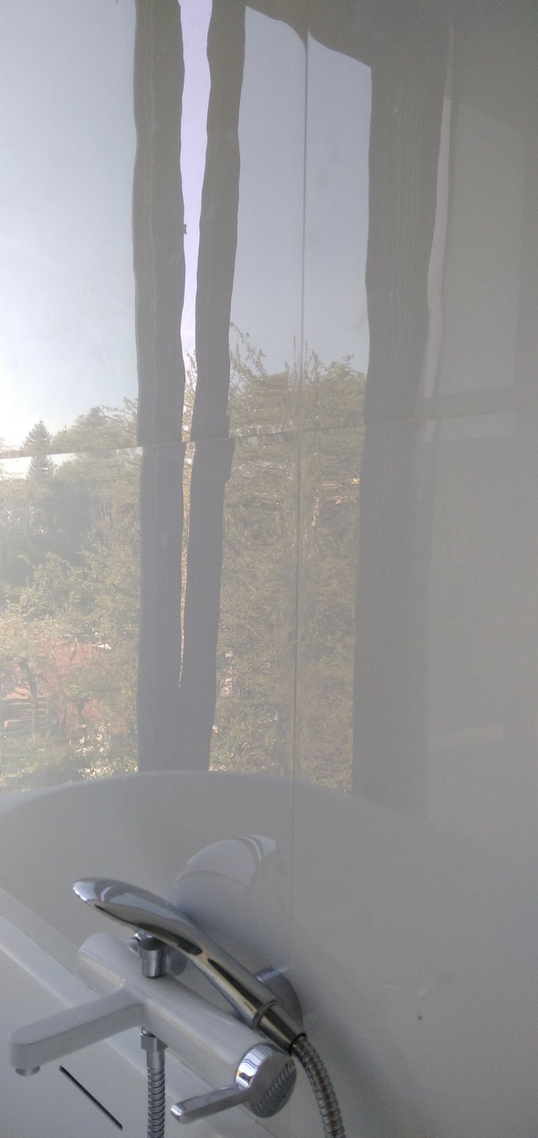 Biała, błyszcząca ściana w łazience z widoczną wanną i baterią prysznicową, w płytkach odbija się widok drzew za oknem.