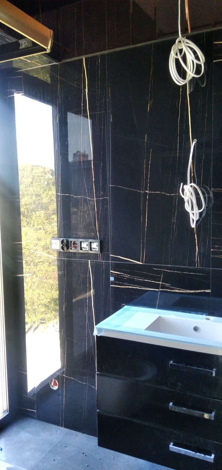 Częściowo wykończona łazienka z czarnymi płytkami imitującymi marmur ze złotymi żyłami, wisząca biała umywalka z czarną szafką, widoczne kable i otwór w ścianie, okno z widokiem na zieleń.