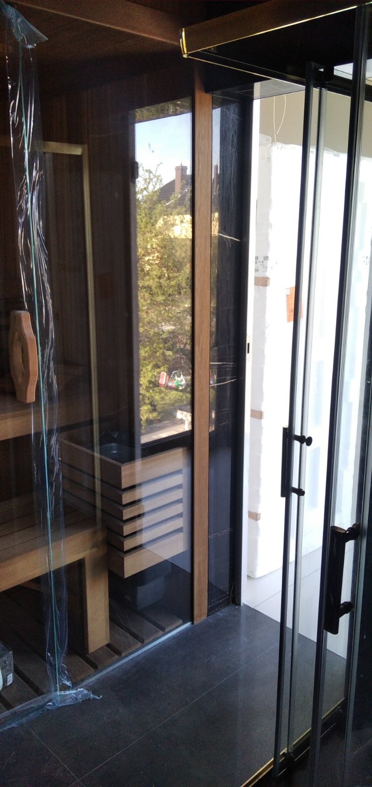 Nowoczesna sauna z drewnianym wykończeniem i szklanymi drzwiami przesuwnymi, widok na zewnątrz przez okno, czarna podłoga z płytek.