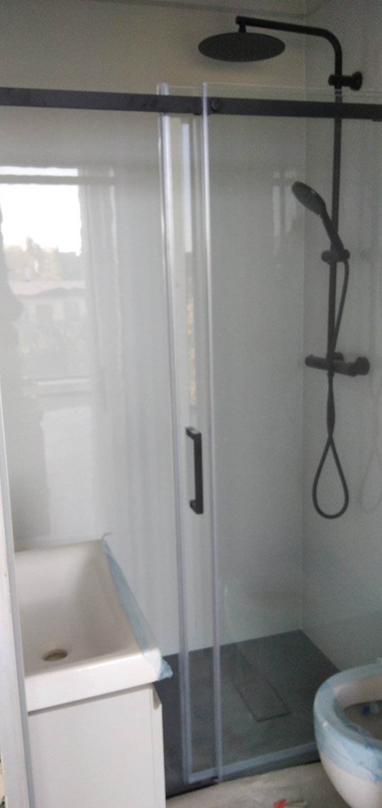 Nowoczesna kabina prysznicowa z czarną armaturą, szklanymi przesuwnymi drzwiami i brodzikiem w odcieniach szarości, widoczna wanna z umywalką i toaleta w folii ochronnej.