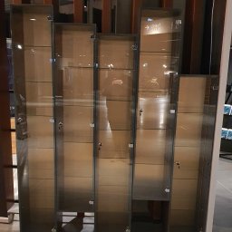 Meble na Wymiar Artur Bisztyga - Szereg szklanych witryn ekspozycyjnych z metalowymi ramami i zamkami, o różnej wysokości, ustawionych na szarym tle podłogi.