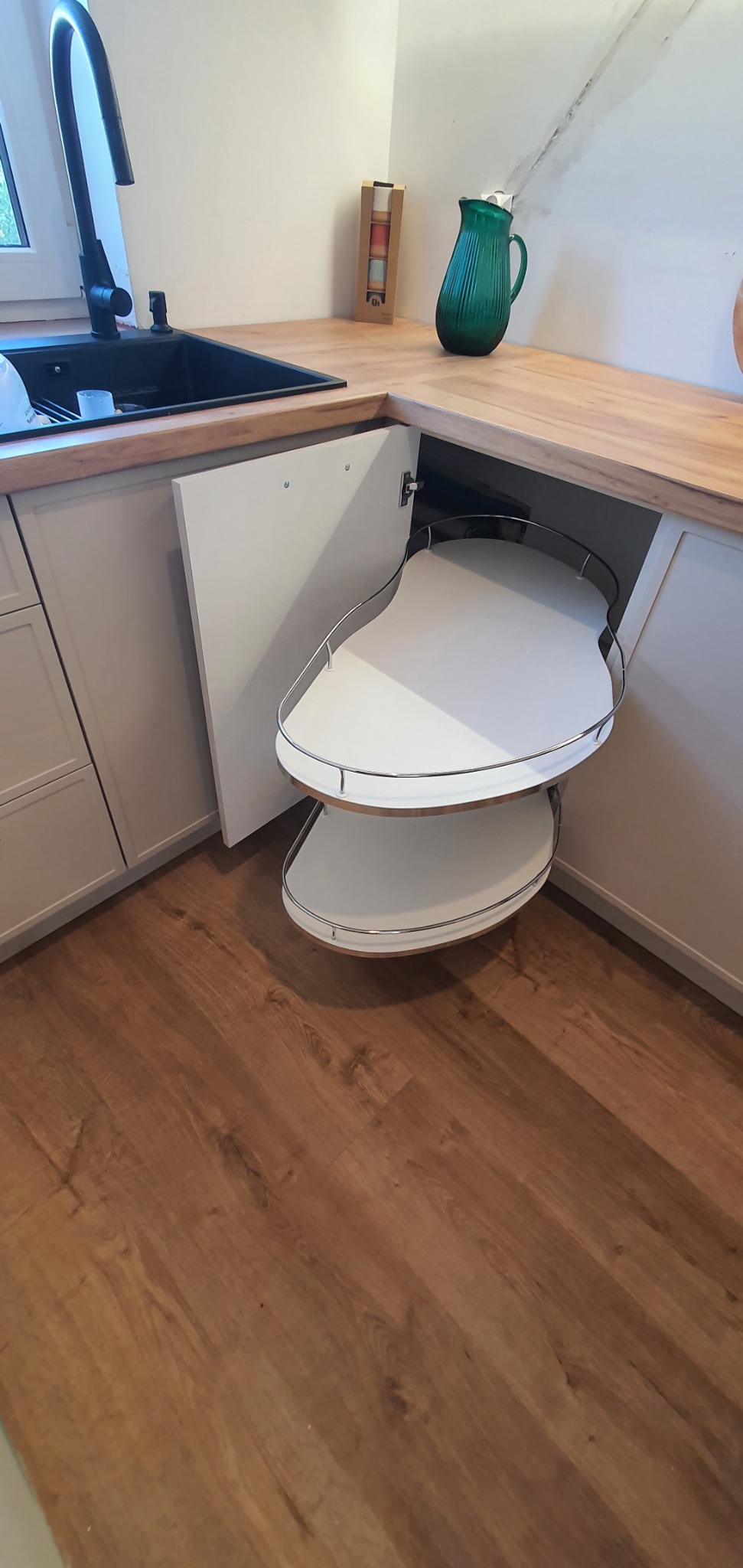 Wnętrze szafki narożnej z obrotowymi półkami w kuchni z jasnymi frontami i drewnianym blatem, widoczny zlew i bateria.