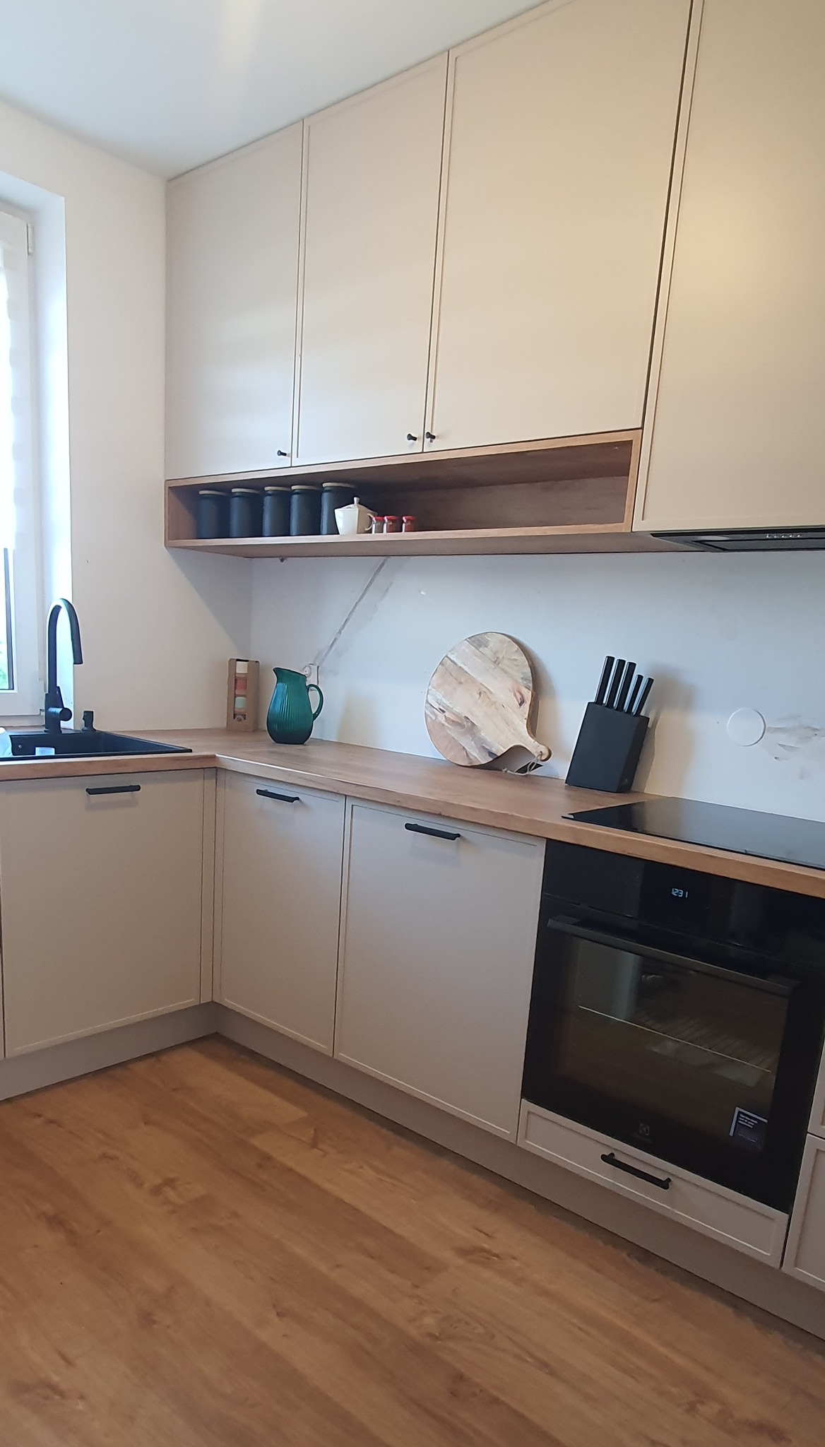 Jasna kuchnia na wymiar z drewnianym blatem, czarnymi uchwytami i otwartą półką z przyprawami, uzupełniona o czarny zlew, deskę do krojenia i zestaw noży.