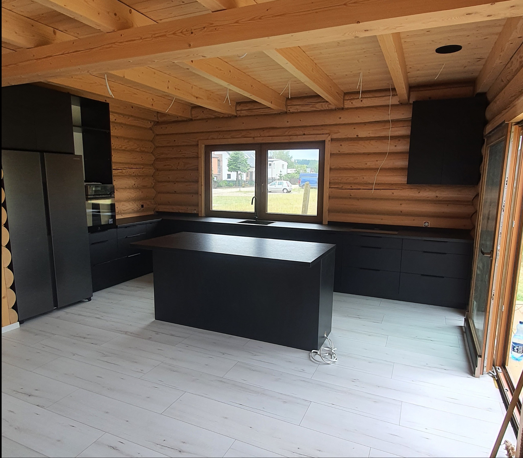 Wnętrze drewnianego domu z czarną, nowoczesną zabudową kuchenną z wyspą, widok na okno i białą podłogę.