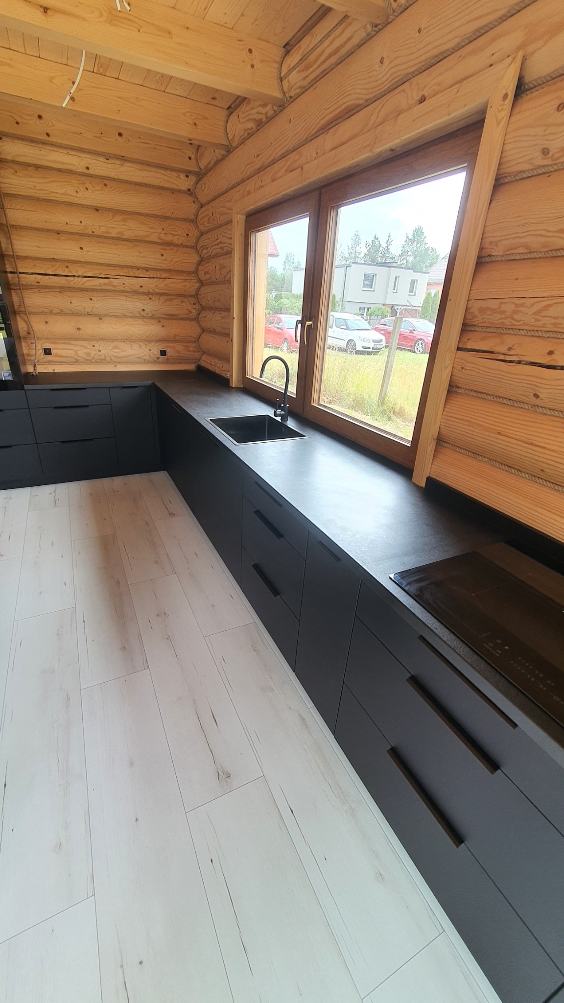 Wnętrze kuchni w drewnianym domu z czarnymi, matowymi szafkami, blatem i zlewozmywakiem przy oknie z widokiem na podwórko, jasna podłoga imitująca drewno.