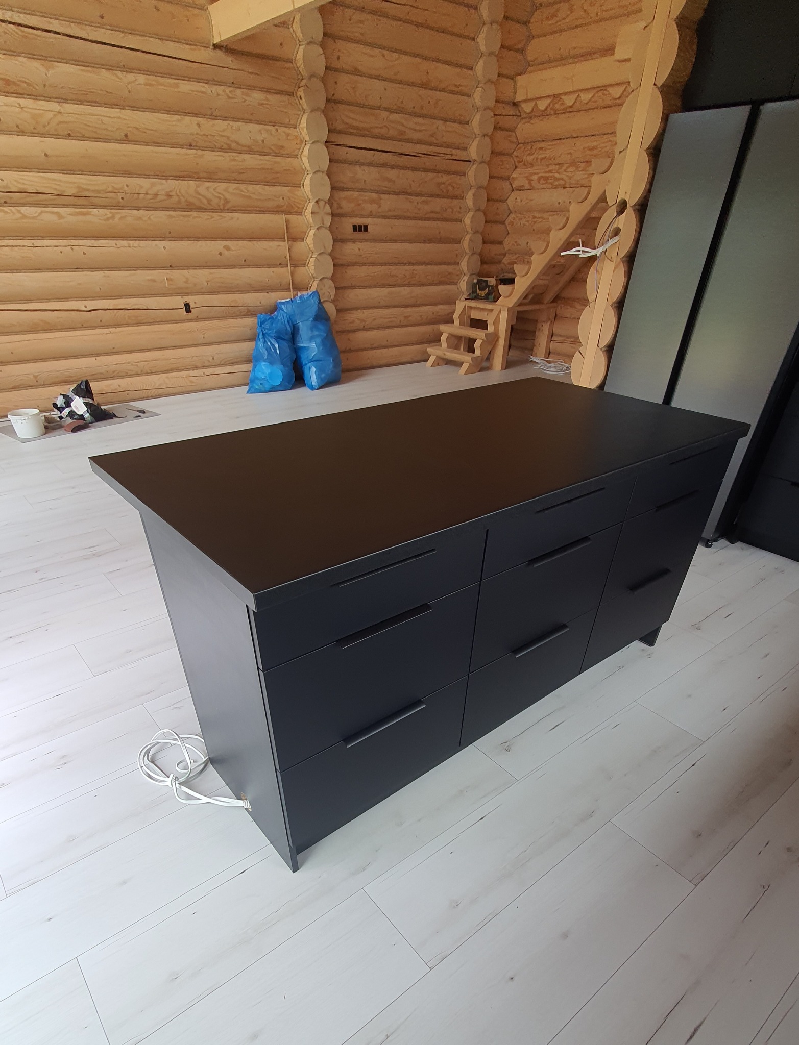 Czarna wyspa kuchenna z szufladami, w surowym wnętrzu domu z bali w Banino, z widocznymi workami i schodkami w tle.